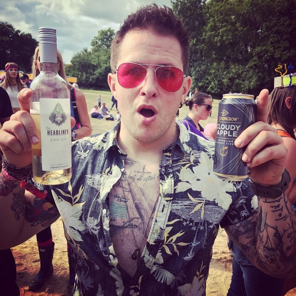 Rob holding a can of cider and a glass of wine wearing red tinted sunglasses at Glastonbury Festival